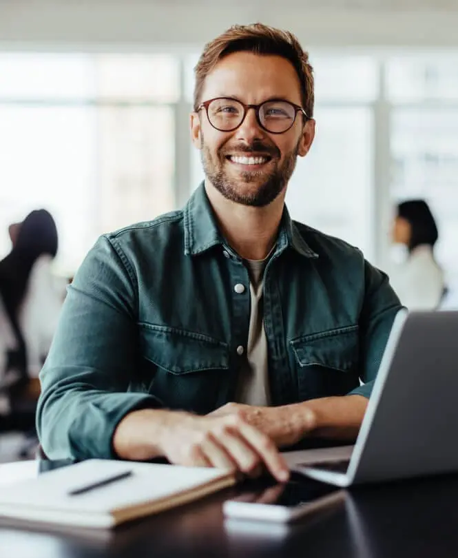 A smiling man at a desk in front of a laptop.