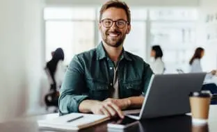 A smiling man at a desk in front of a laptop.