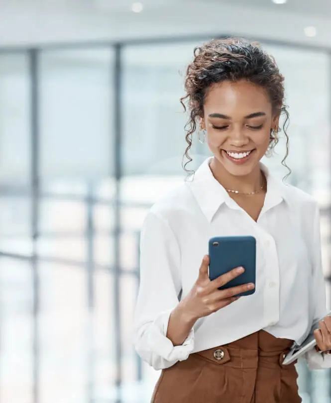 A woman in the office uses a smartphone and holds a tablet.