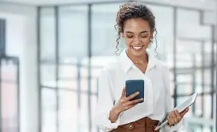 A woman in the office uses a smartphone and holds a tablet.