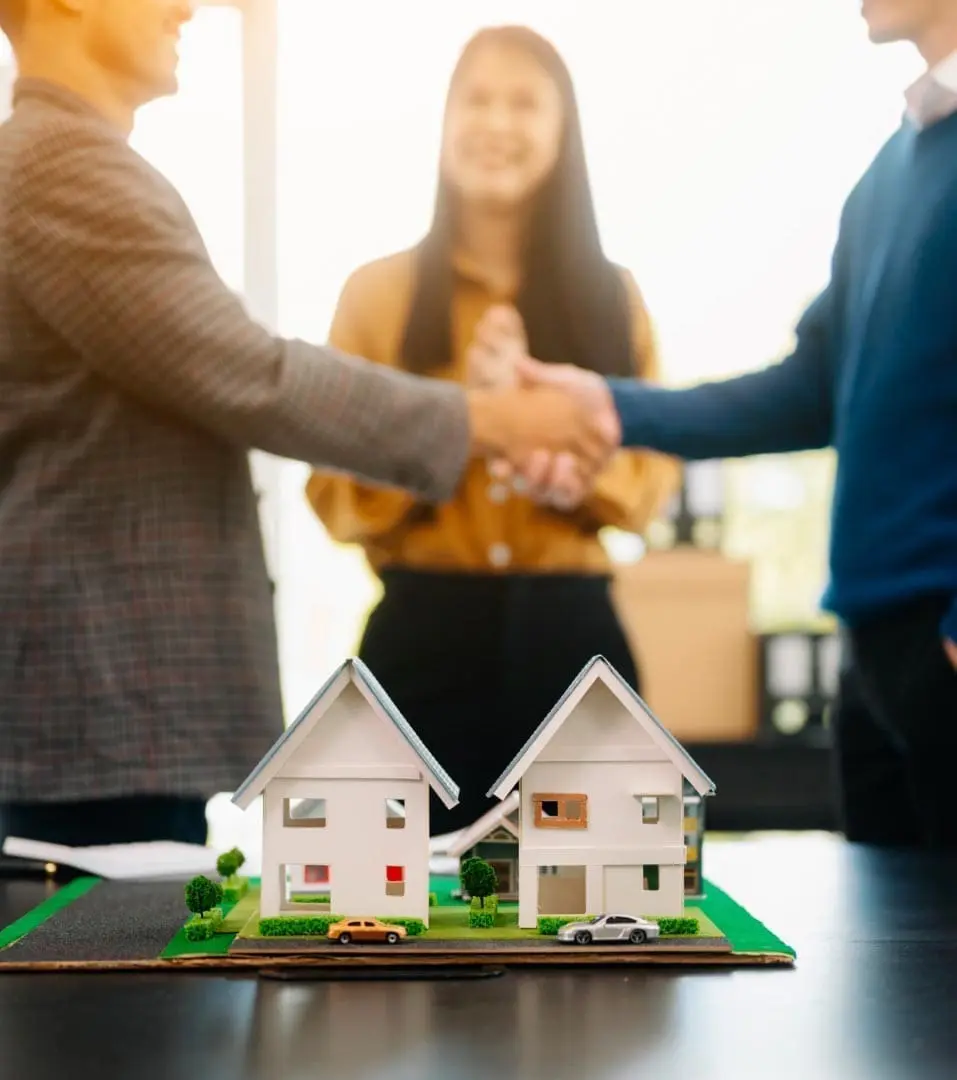 Two people shake hands in front of a real estate agent, with a model of two houses in the foreground.