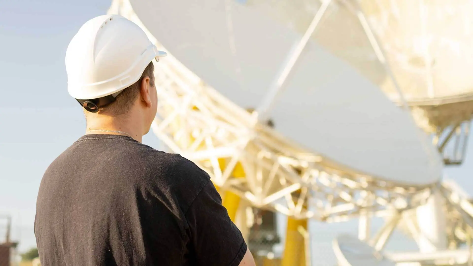 Tecnico con elmetto di spalle di fronte a una grande antenna di telecomunicazioni