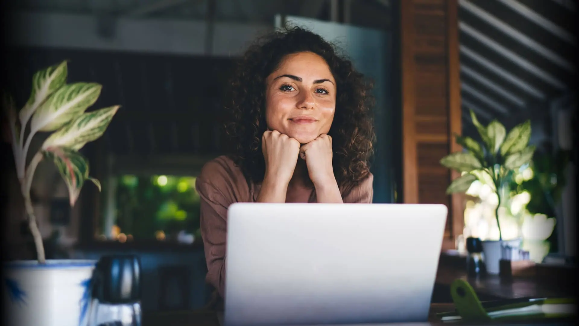 Woman with satisfied expression, at the desk behind a laptop, her face resting on her arms with closed fists.