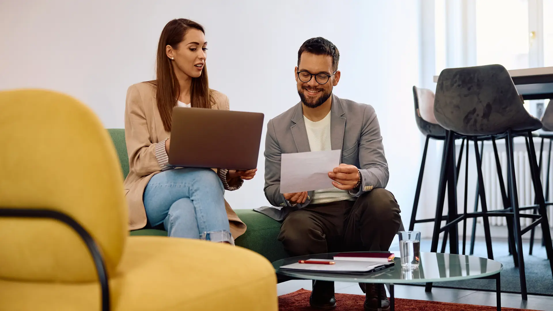 A woman and a man sitting on a sofa looking at a laptop and a paper