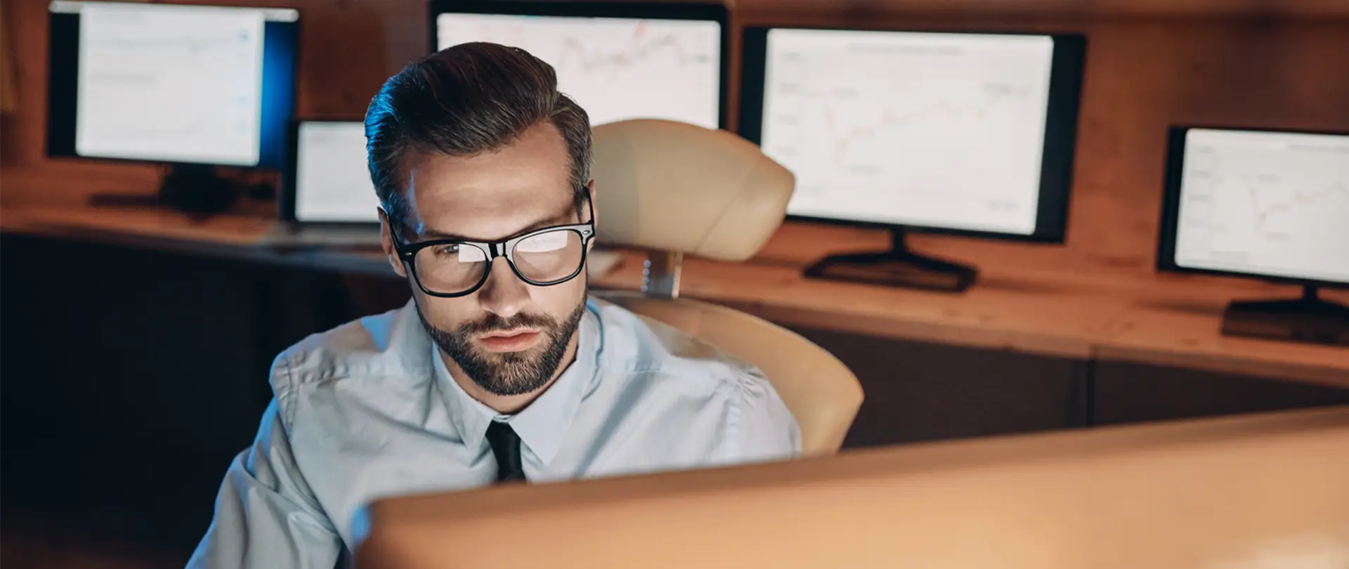 Man in glasses and elegant suit busy working in a room with many monitors.